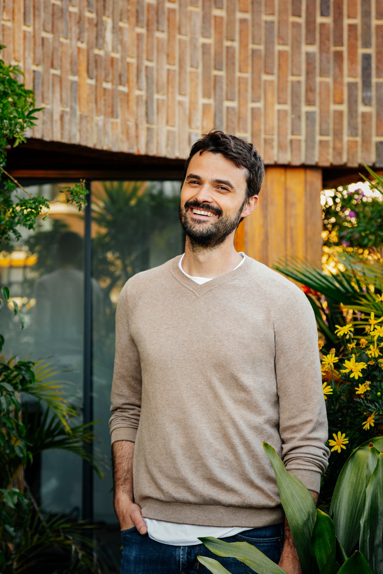 Man smiling in front of brick building with plants and yellow flowers.