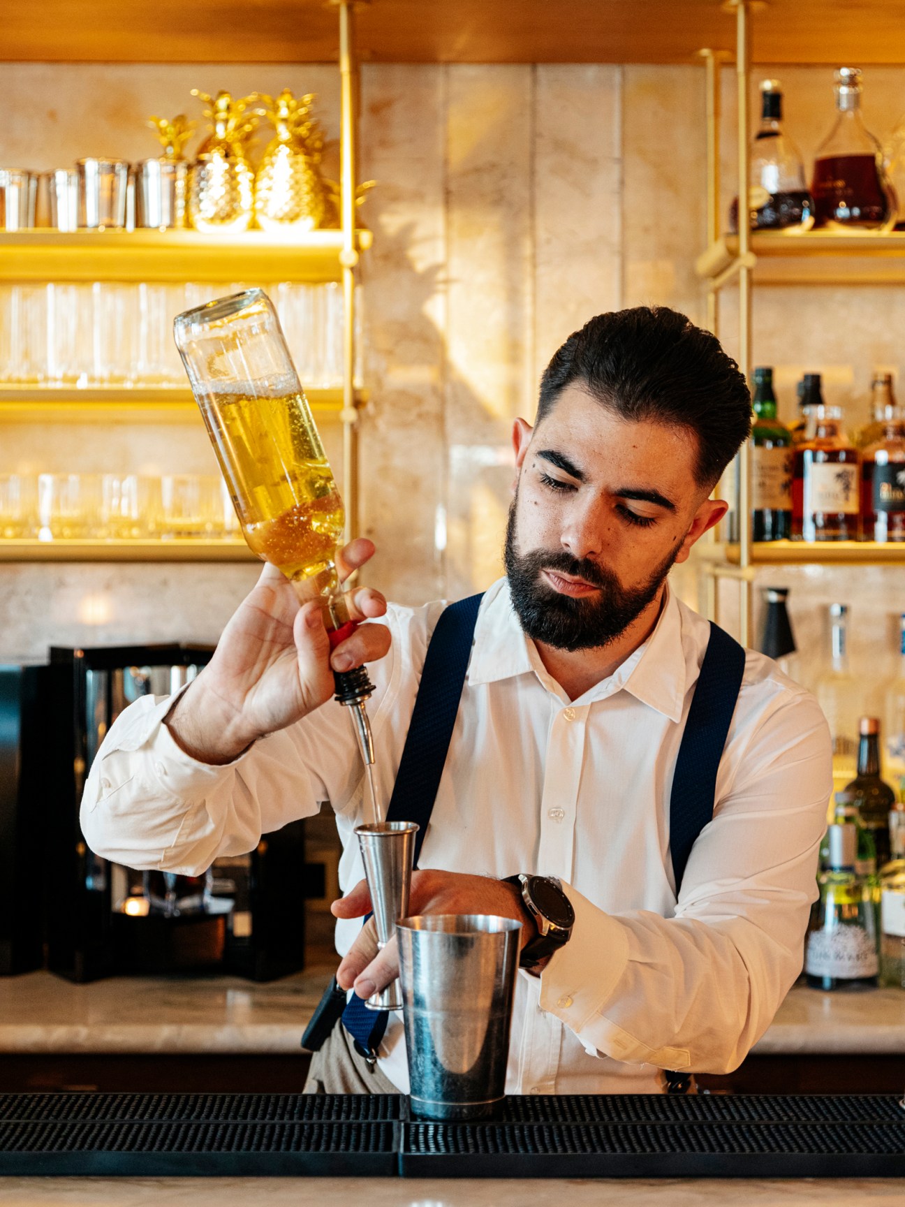 Professional bartender in white shirt and suspenders pouring golden liquid from bottle into metal cocktail shaker