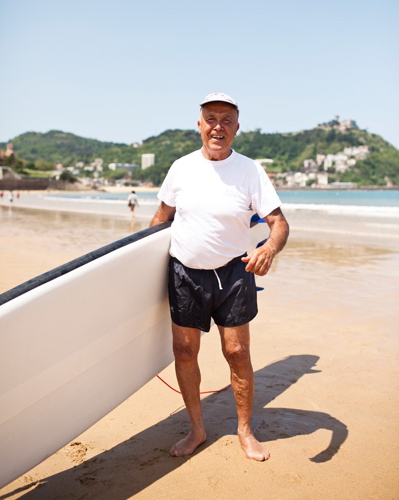 Man holding a surfboard on a sandy beach with hillside buildings in the background.
