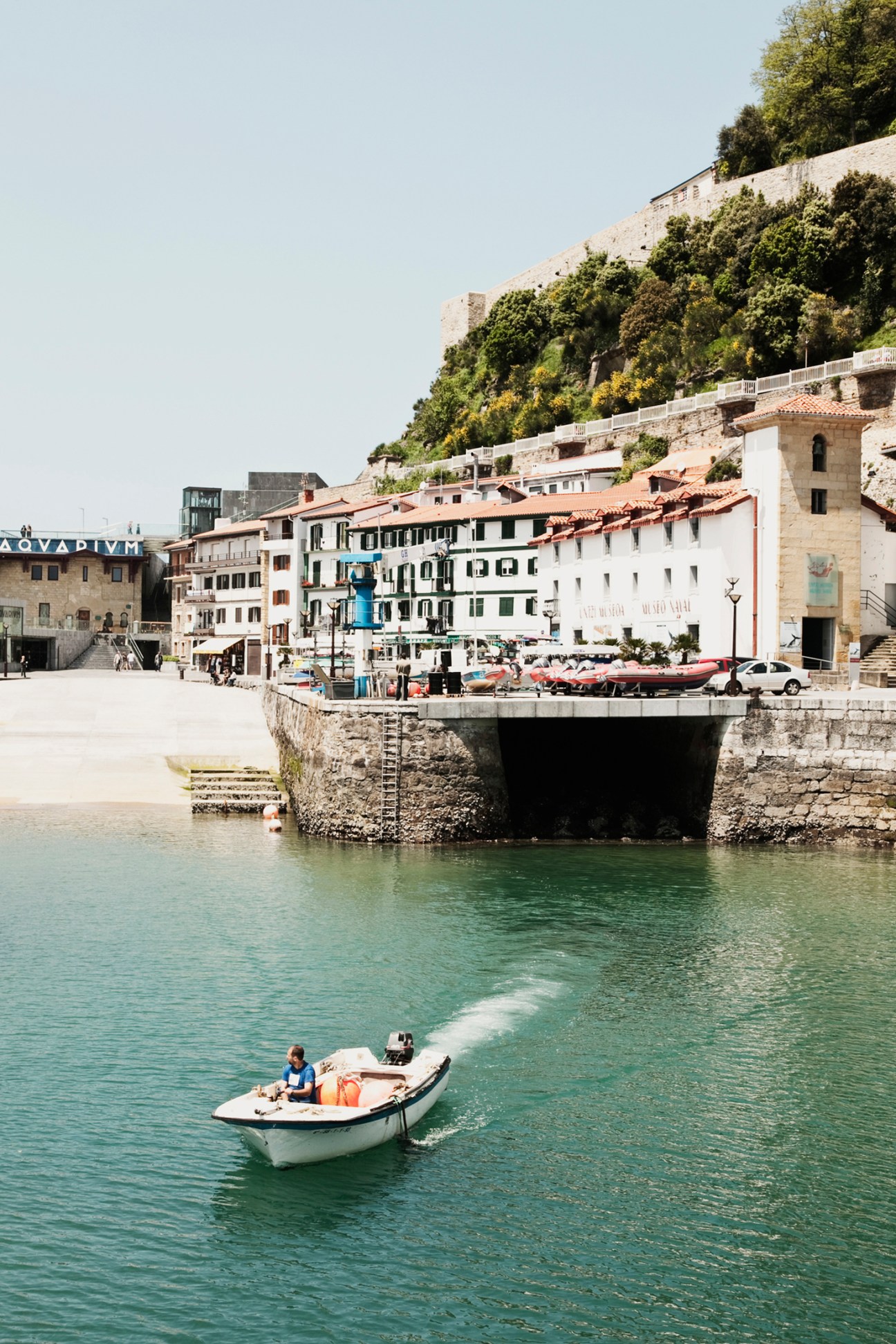 A small white boat on turquoise water in front of historic stone buildings and a fortified cliff in a Mediterranean coastal town.