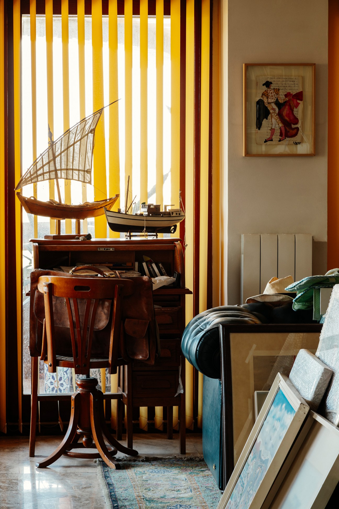 Interior of a residential space featuring a wooden desk with model sailing ships near striped yellow and white curtains, a framed historical illustration on the beige wall, and stacked artwork leaning against furniture in the foreground