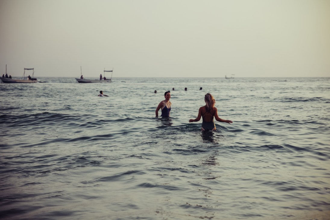 Swimmers in the sea with traditional fishing boats anchored in the background.