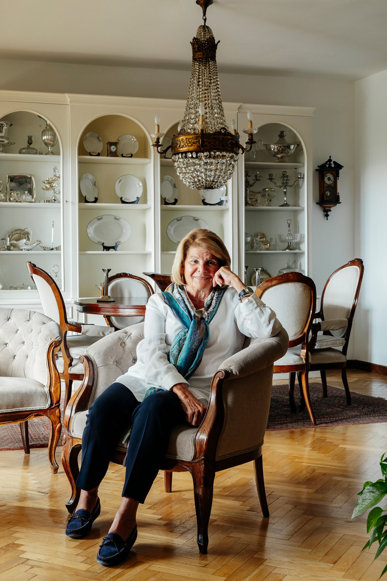 María Adela sitting in her elegantly furnished apartment with crystal chandelier and china display cabinets