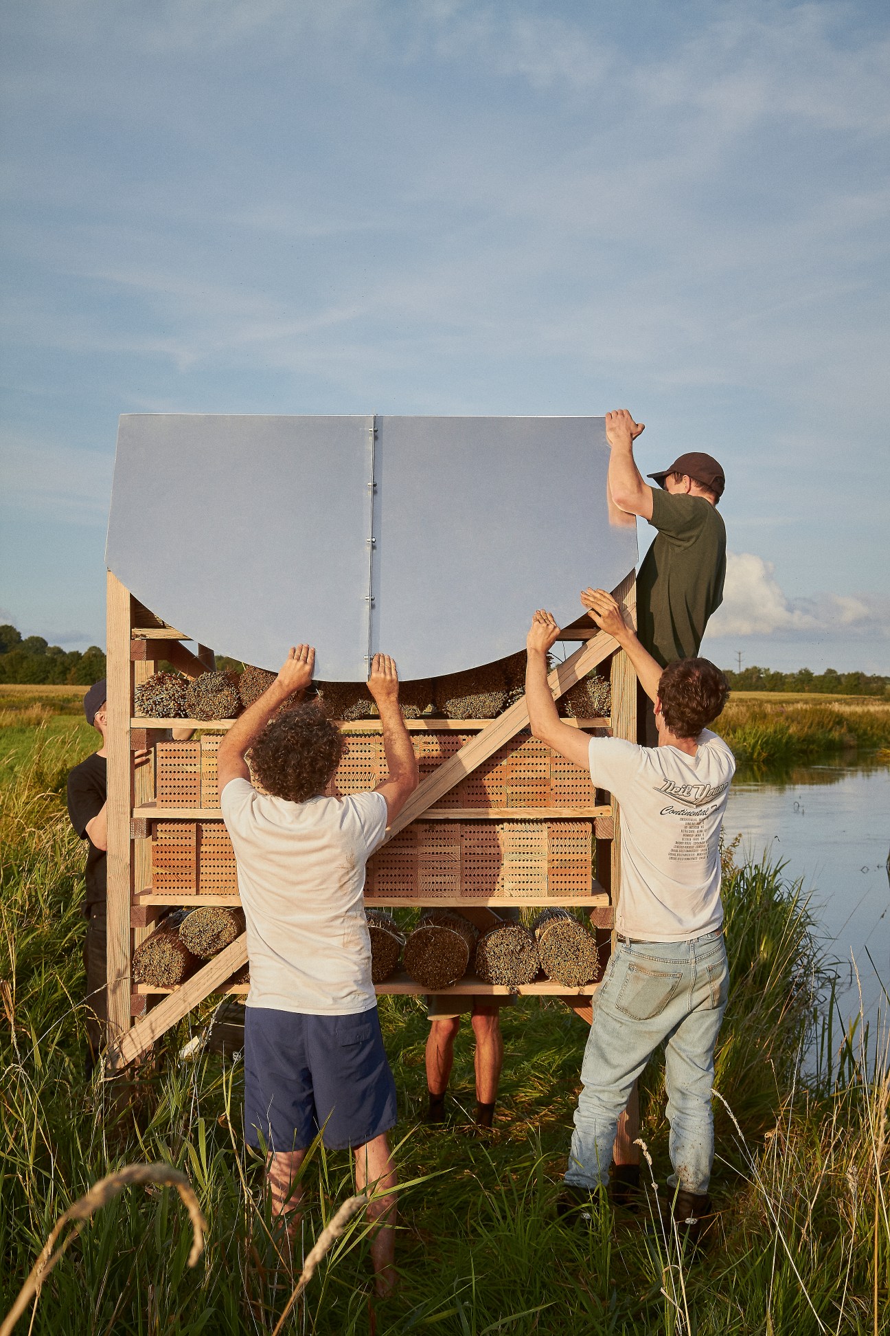 Three people assembling a wooden beehive structure in a grassy field near water