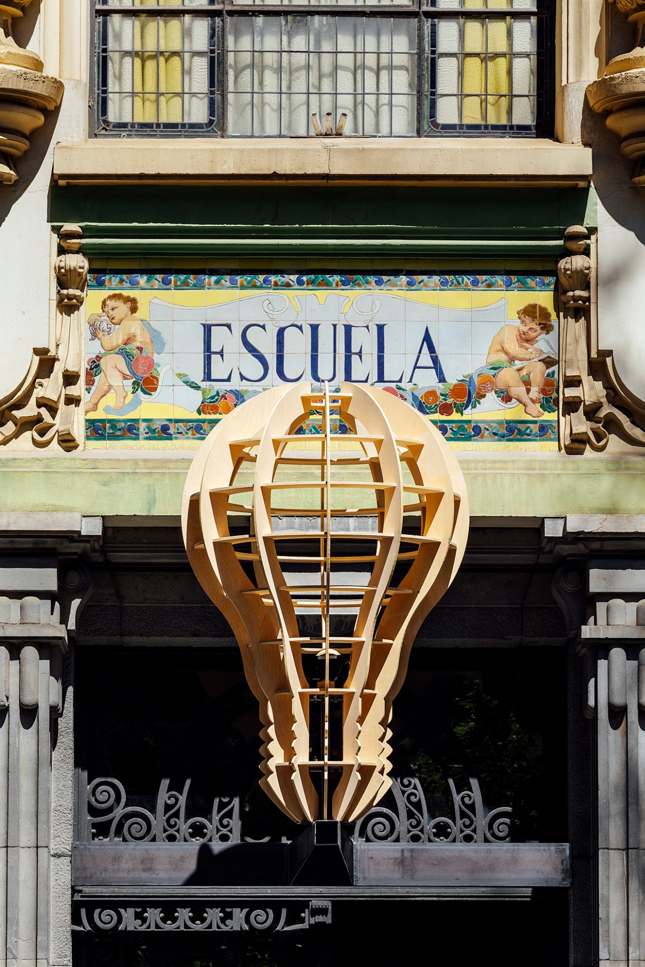 Large wooden lightbulb-shaped structure suspended above a balcony with 'ESCUELA' tile sign on historic building facade