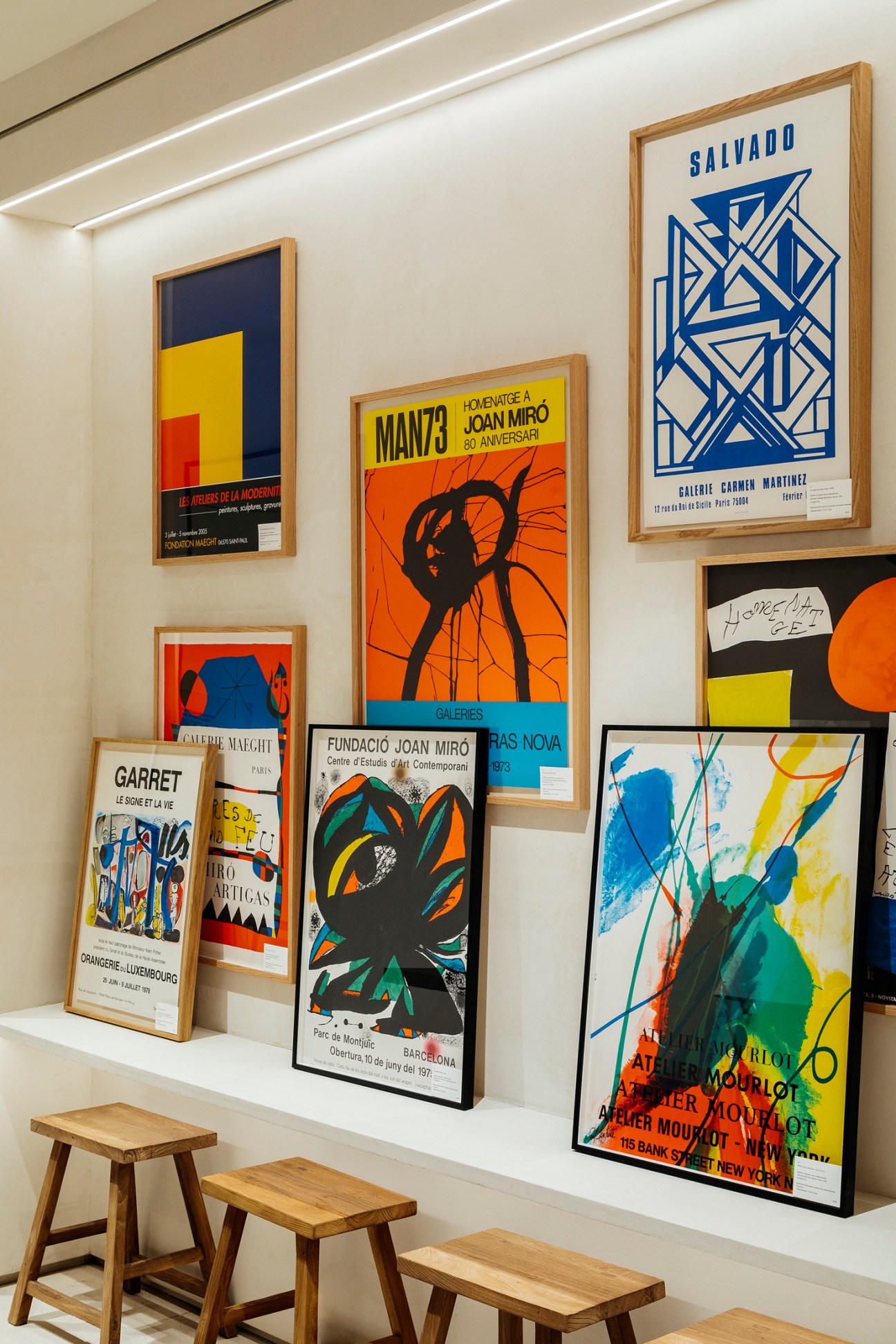 Collection of colorful framed art prints and exhibition posters displayed on white wall above wooden stools