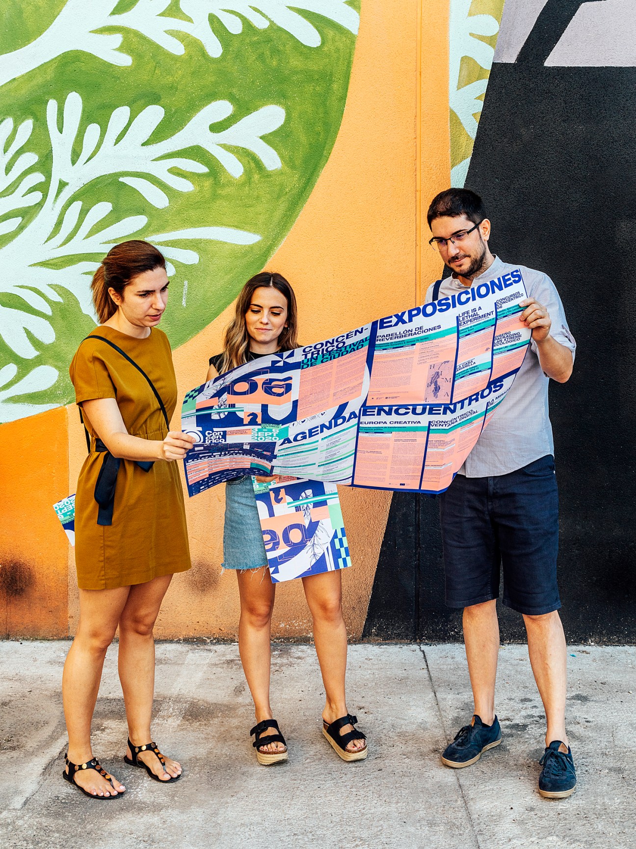 Three people holding and reading colorful festival posters against a vibrant mural wall