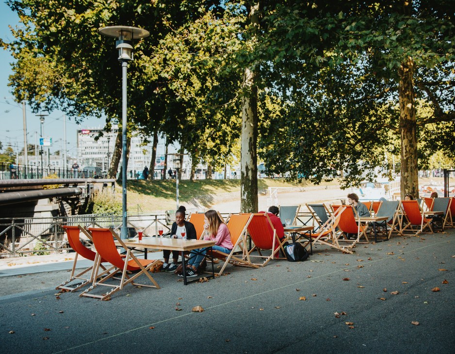 Outdoor seating area with red and orange canvas deck chairs under tall leafy trees beside a waterway in Nantes.