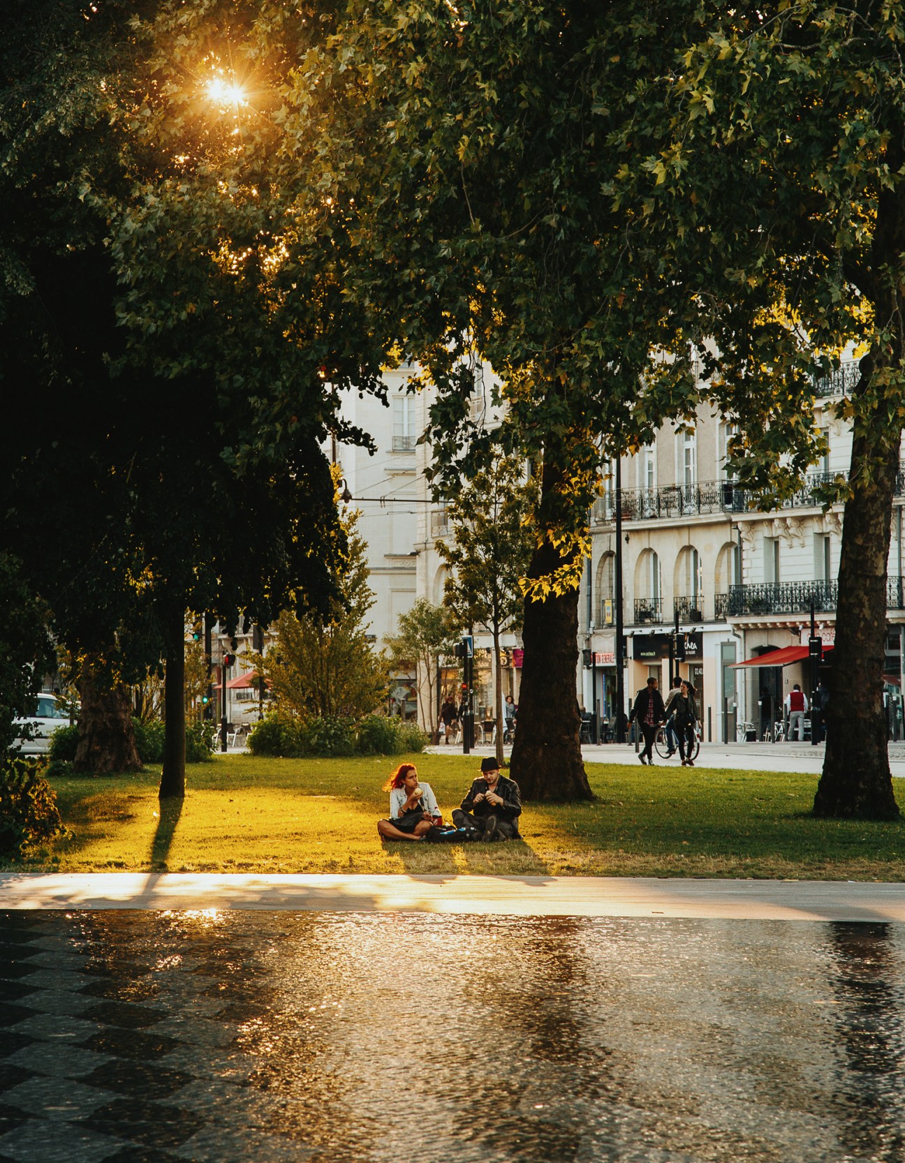 Two people sitting beneath shaded trees in a Nantes city square with historic buildings and storefronts visible in the background.
