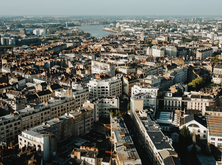 Aerial view of Nantes cityscape with terracotta roofs, the Loire River, and surrounding urban sprawl under clear sky.