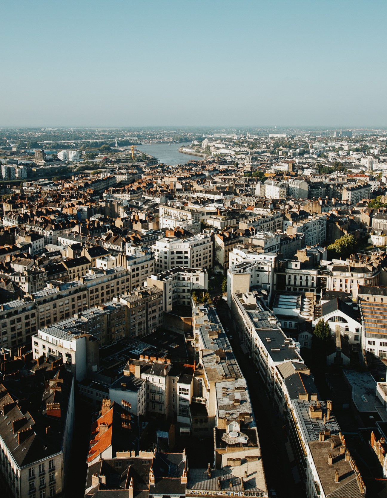 Aerial view of Nantes cityscape with terracotta roofs, the Loire River, and surrounding urban sprawl under clear sky.