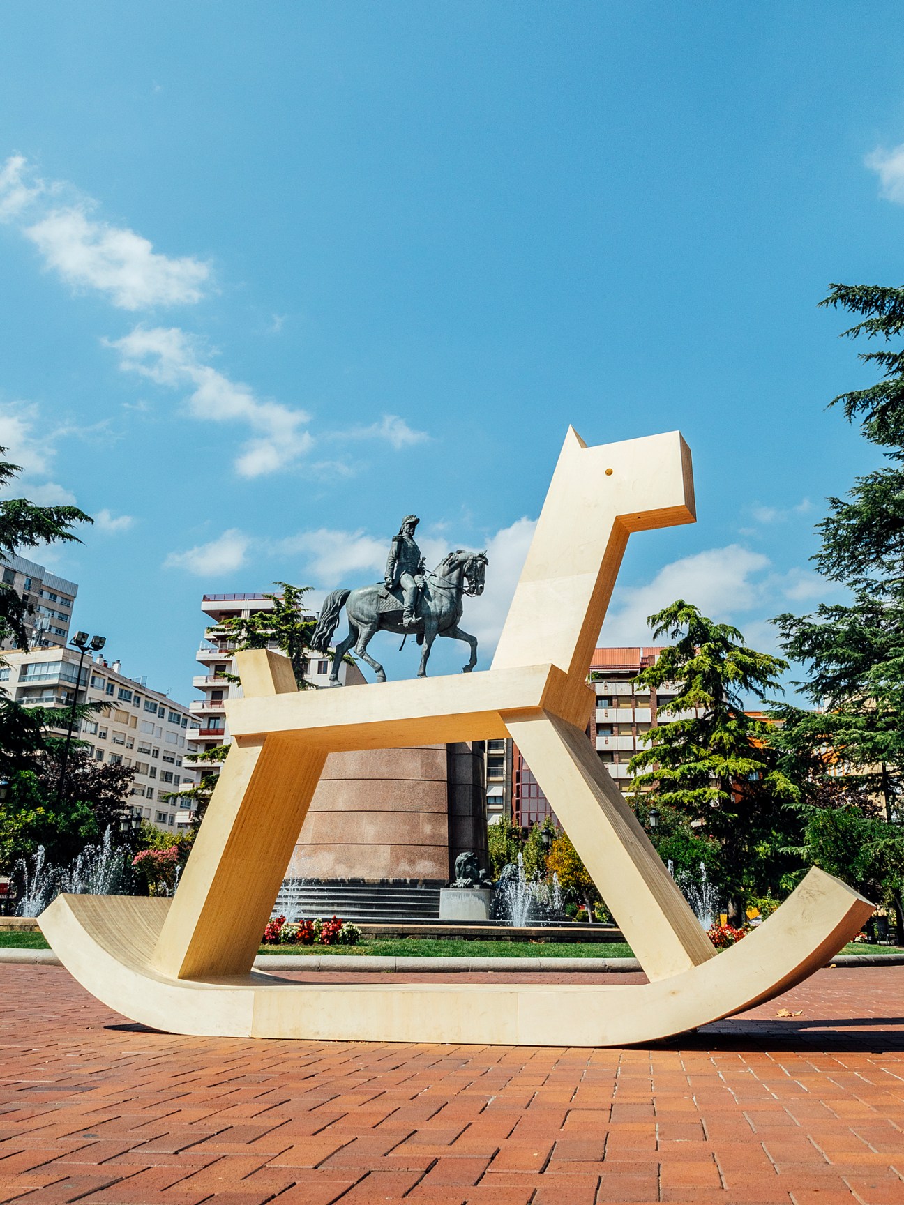 Giant wooden rocking horse sculpture by Iza Rutkowska in a tree-lined square with equestrian statue in background
