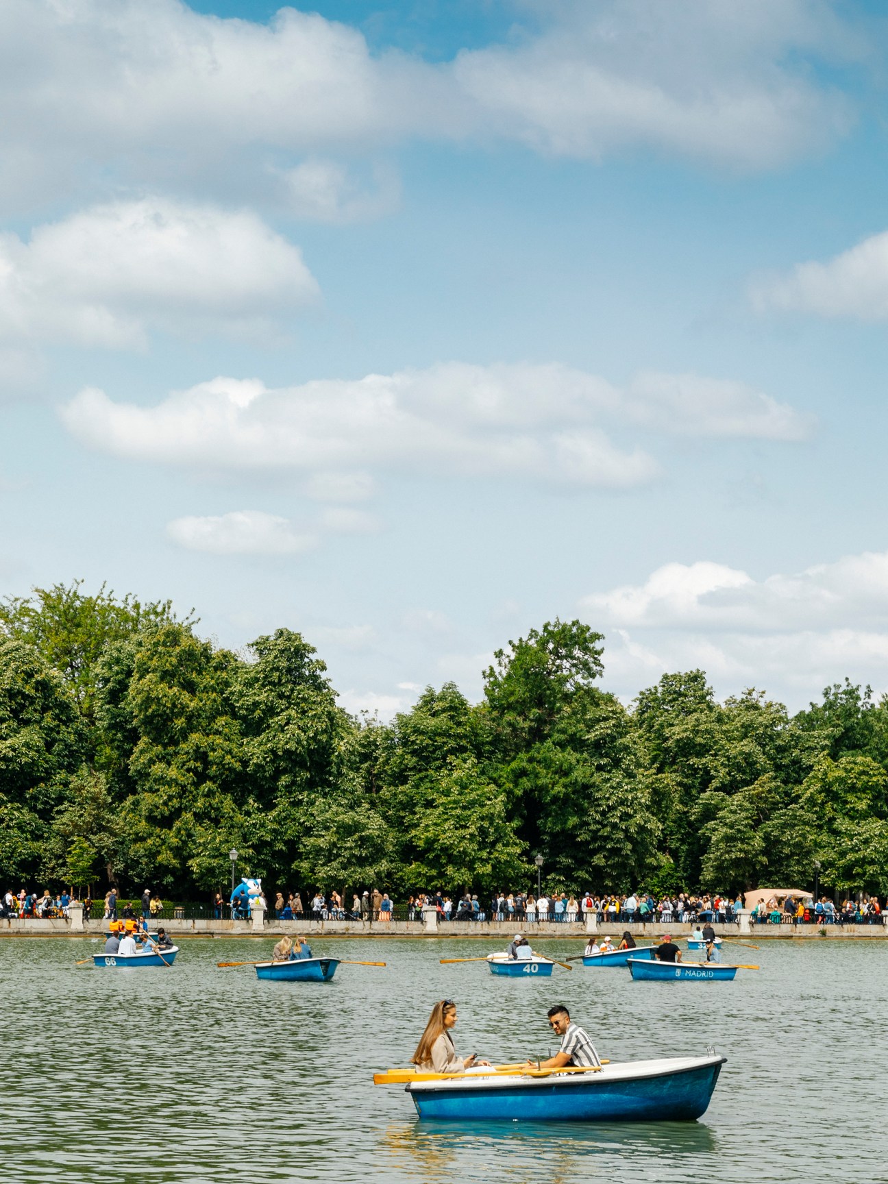 People relaxing in rowboats on a lake with trees and crowds along the shoreline under a partly cloudy sky