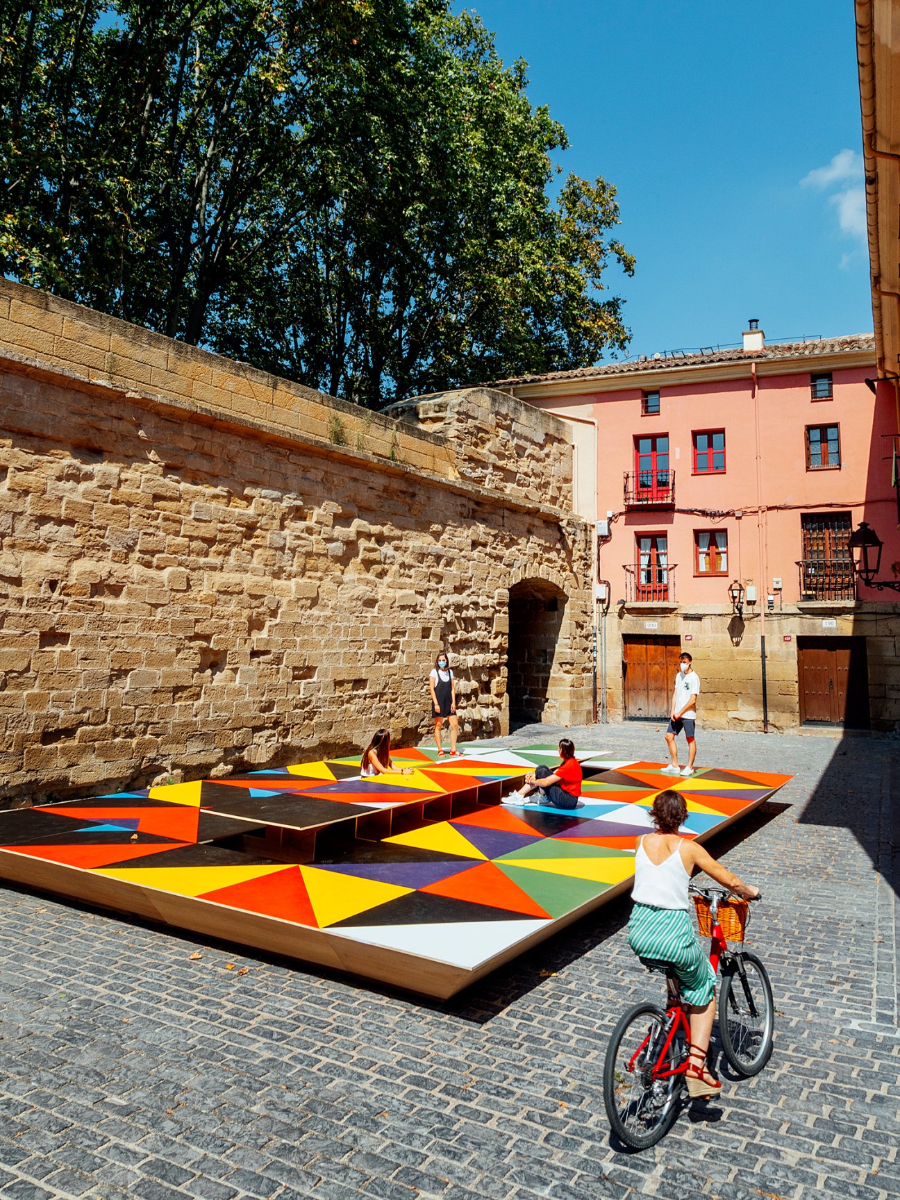 Colorful geometric pavilions in a courtyard beside a historic stone wall and pink building in Belloch, Catalonia.