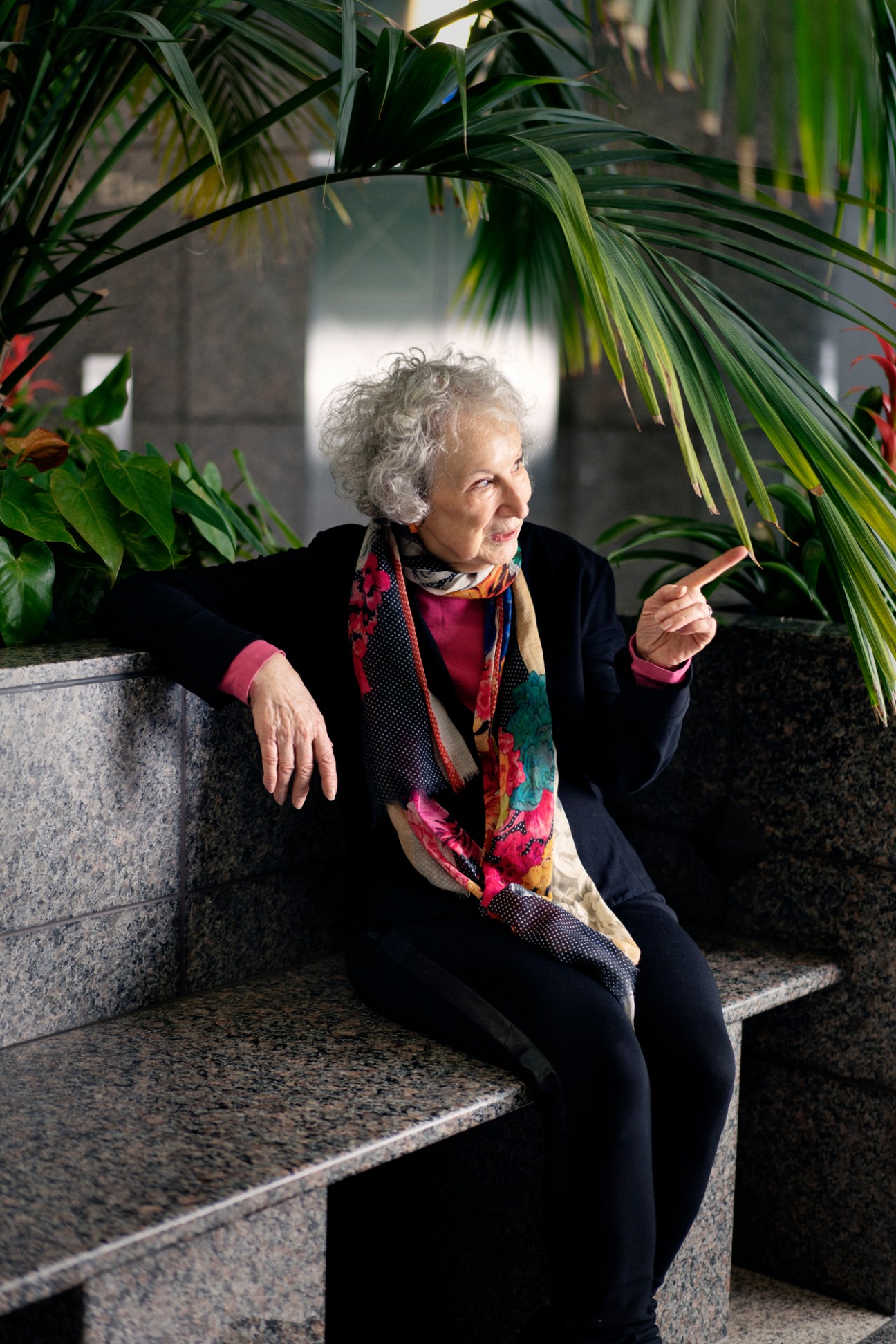 Margaret Atwood sitting on stone steps surrounded by palm plants, wearing a colorful scarf and dark clothing