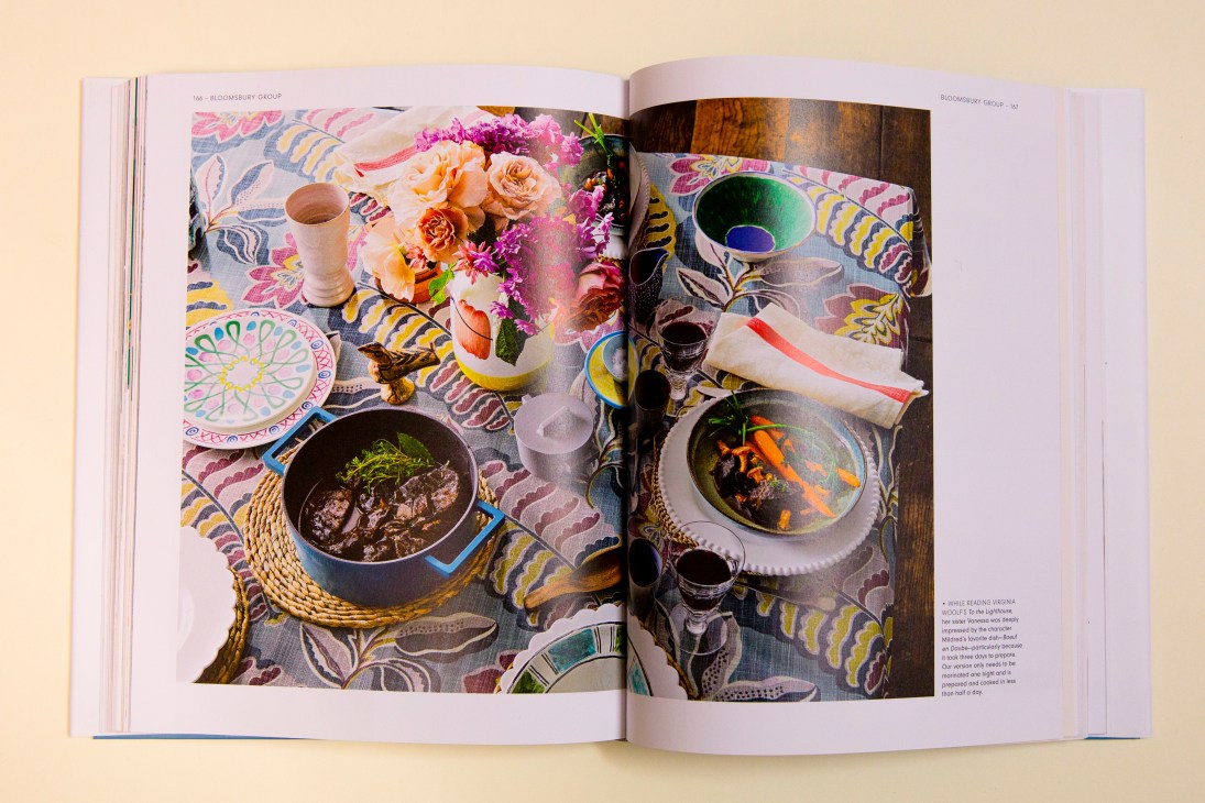 Open cookbook showing colorful food photography with dishes, flowers, and tableware on patterned fabric