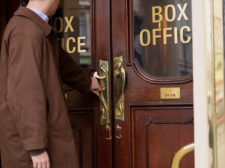 A man in a brown coat opening the glass doors to a theatre box office