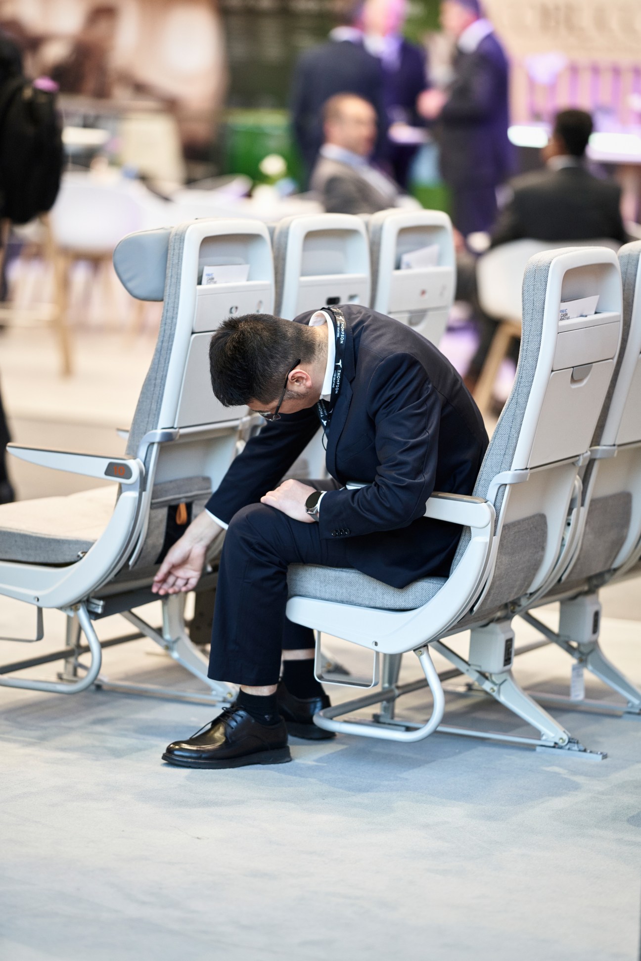 Man in dark business suit sitting hunched over in airline seat at aircraft interiors trade show