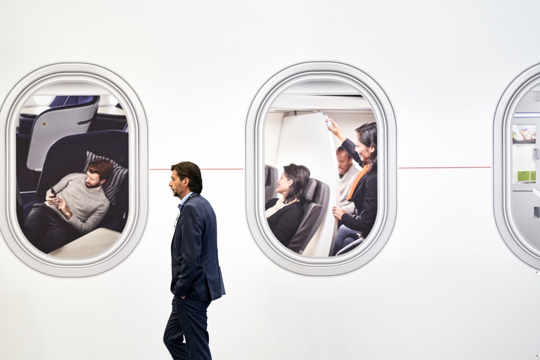 Man in suit walking past aircraft windows showing passengers using in-flight entertainment systems at trade expo