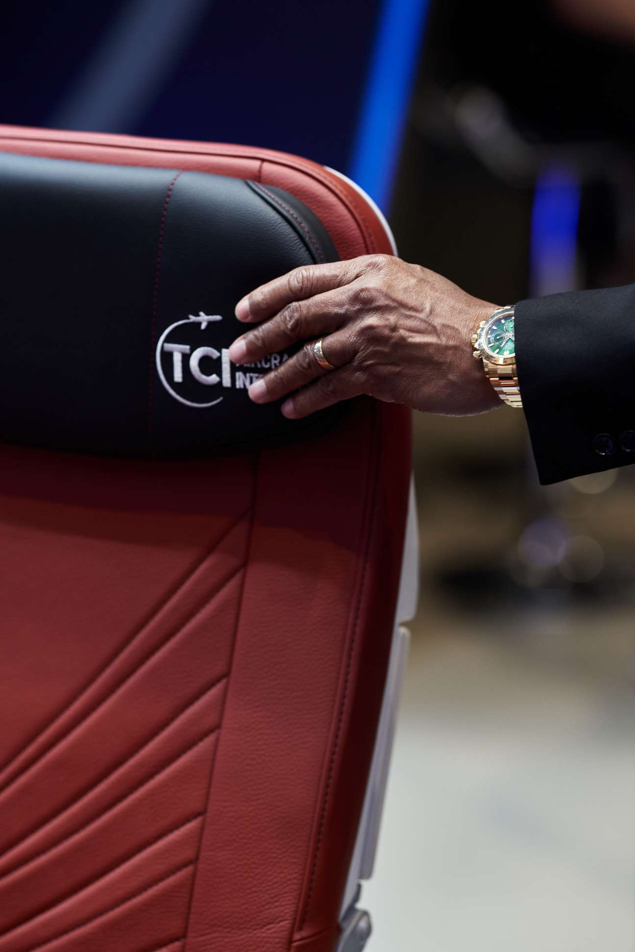 A hand wearing a luxury gold watch rests on a red leather aircraft seat with TCM logo visible
