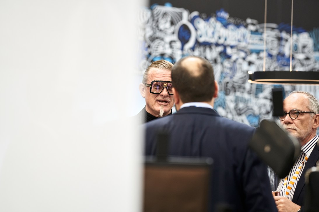 Man in sunglasses speaks to group at aircraft interiors trade show with aviation displays in background