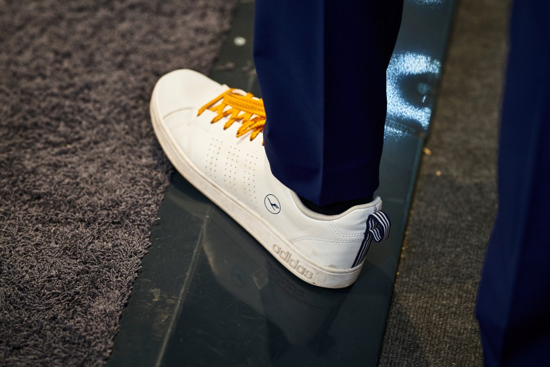Close-up of white Adidas sneaker with orange laces on aircraft cabin floor