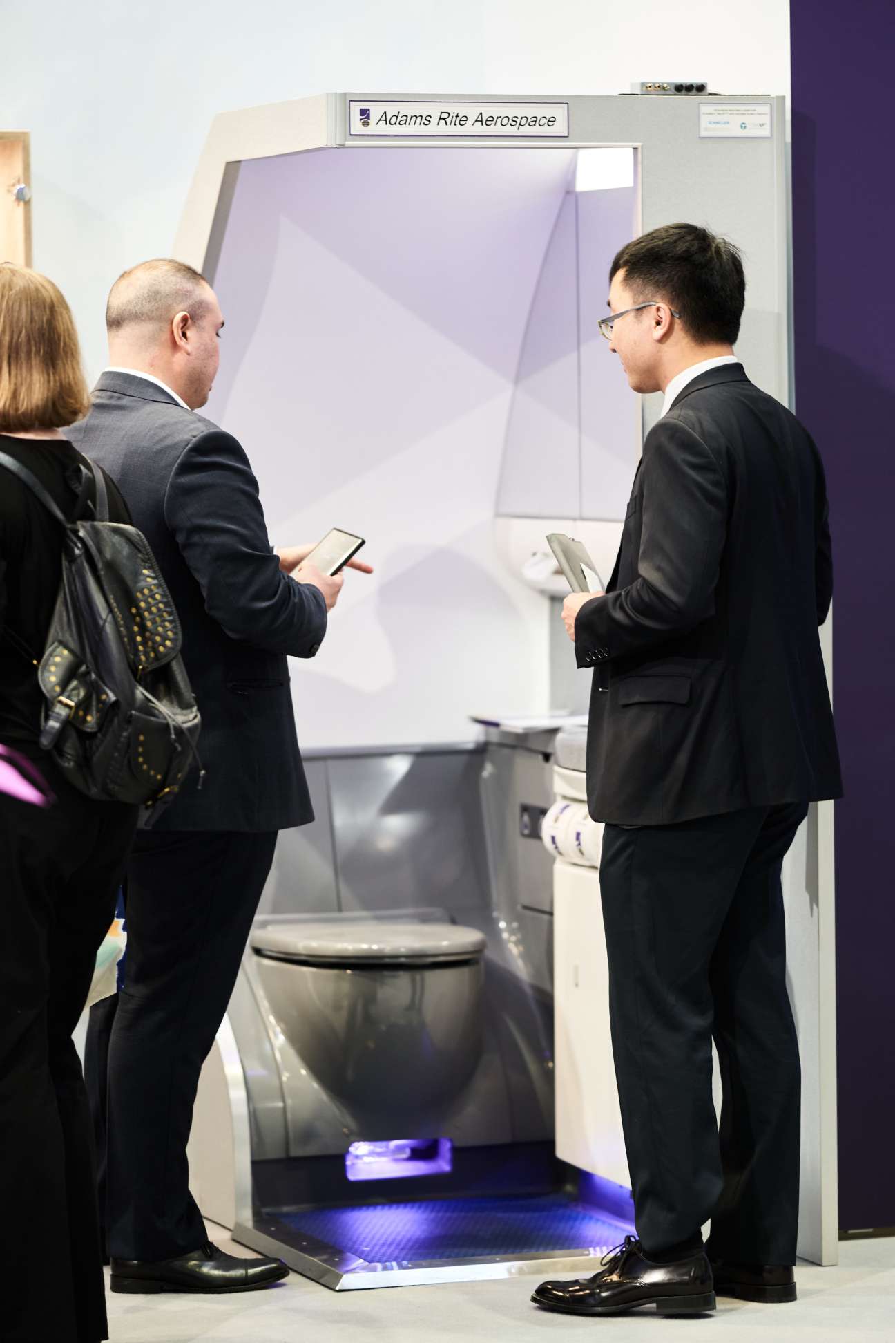 Three business professionals examining an Adams Rite Aerospace 3D printer display at a trade show