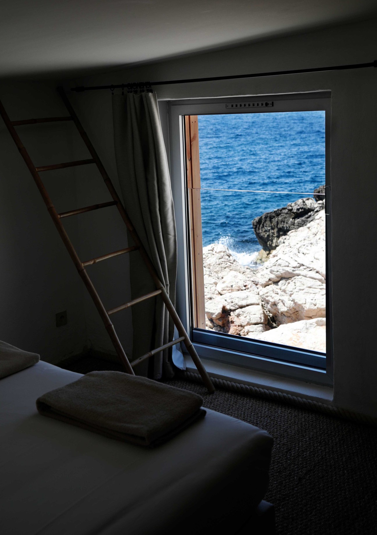Hotel room interior with bed, ladder, window overlooking rocky Mediterranean coastline and blue sea