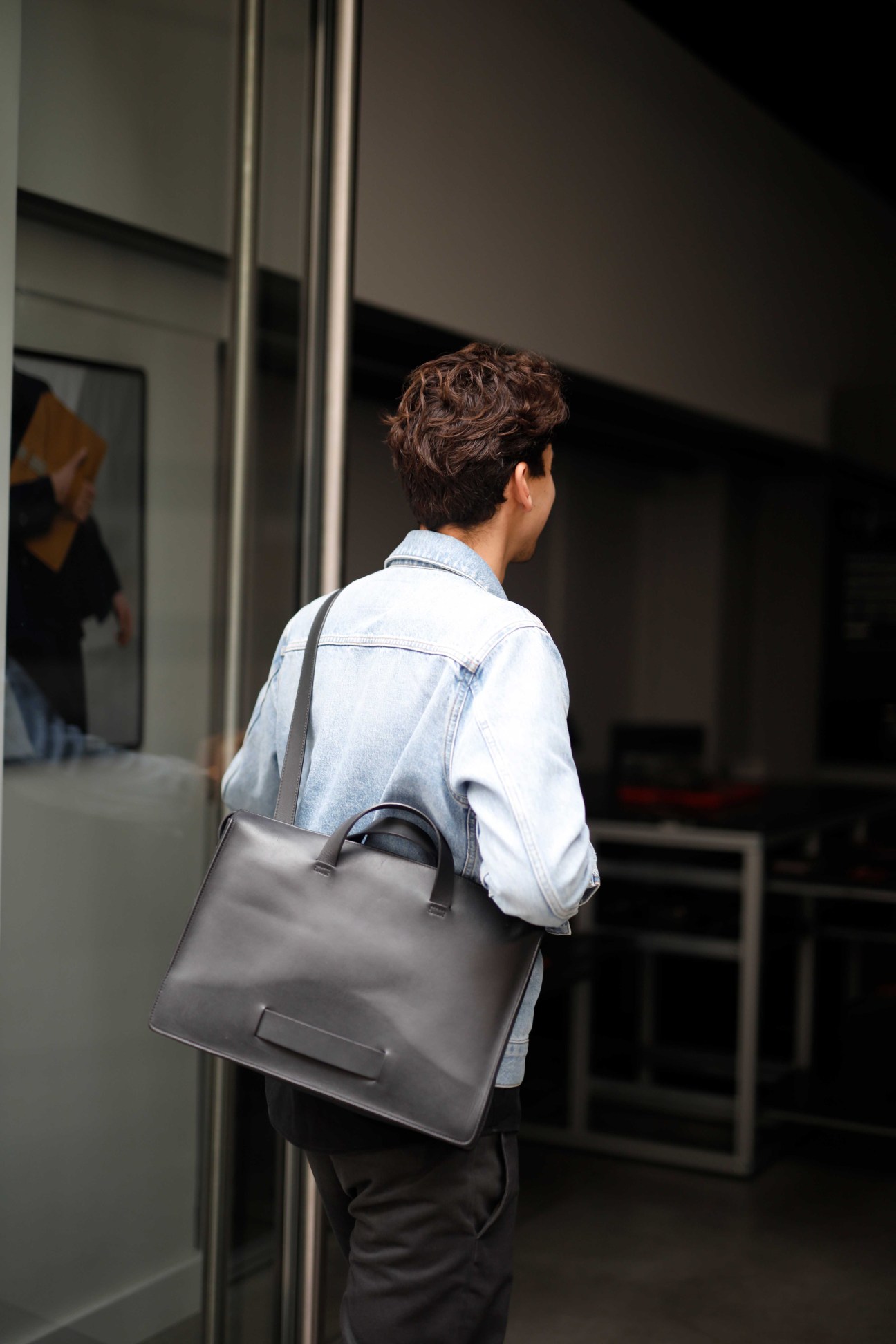 Man in light blue denim shirt carrying grey leather tote bag entering Le Feuillet boutique.