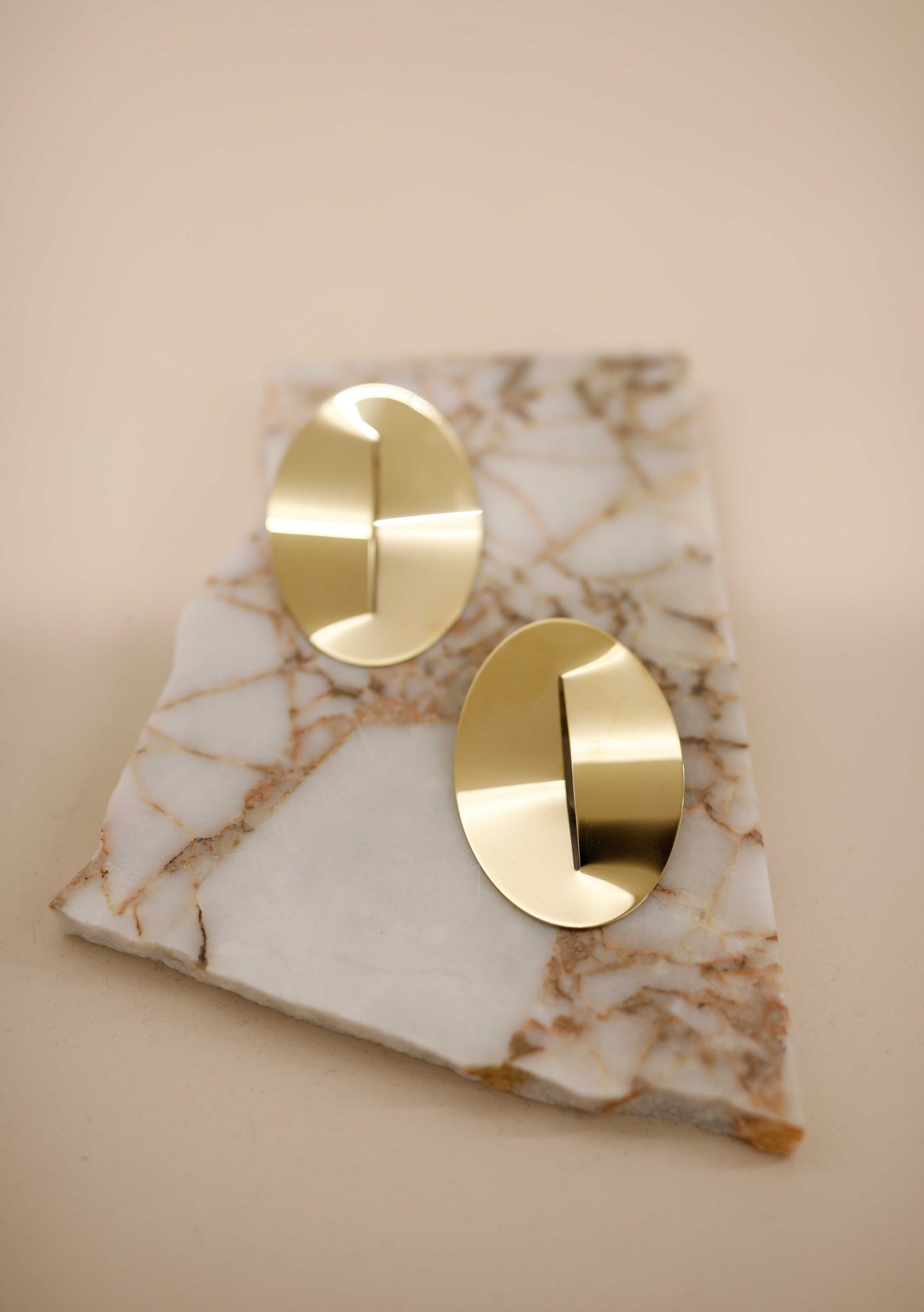 Two gold oval earrings with geometric cutout details displayed on white marble with copper veining.