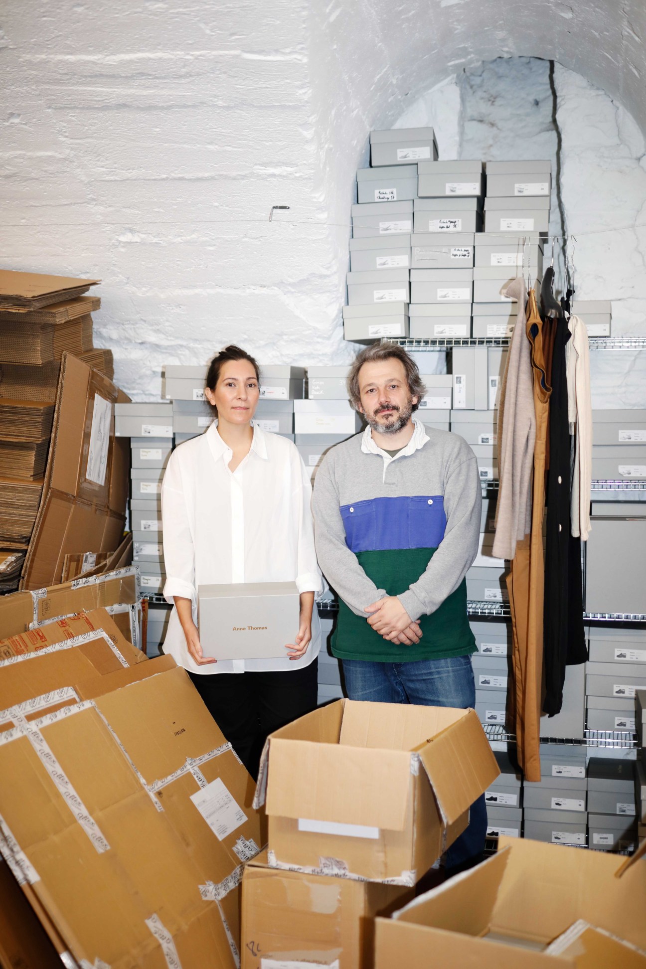 Two people in a warehouse with cardboard boxes and white shelving units stacked with products in the background.