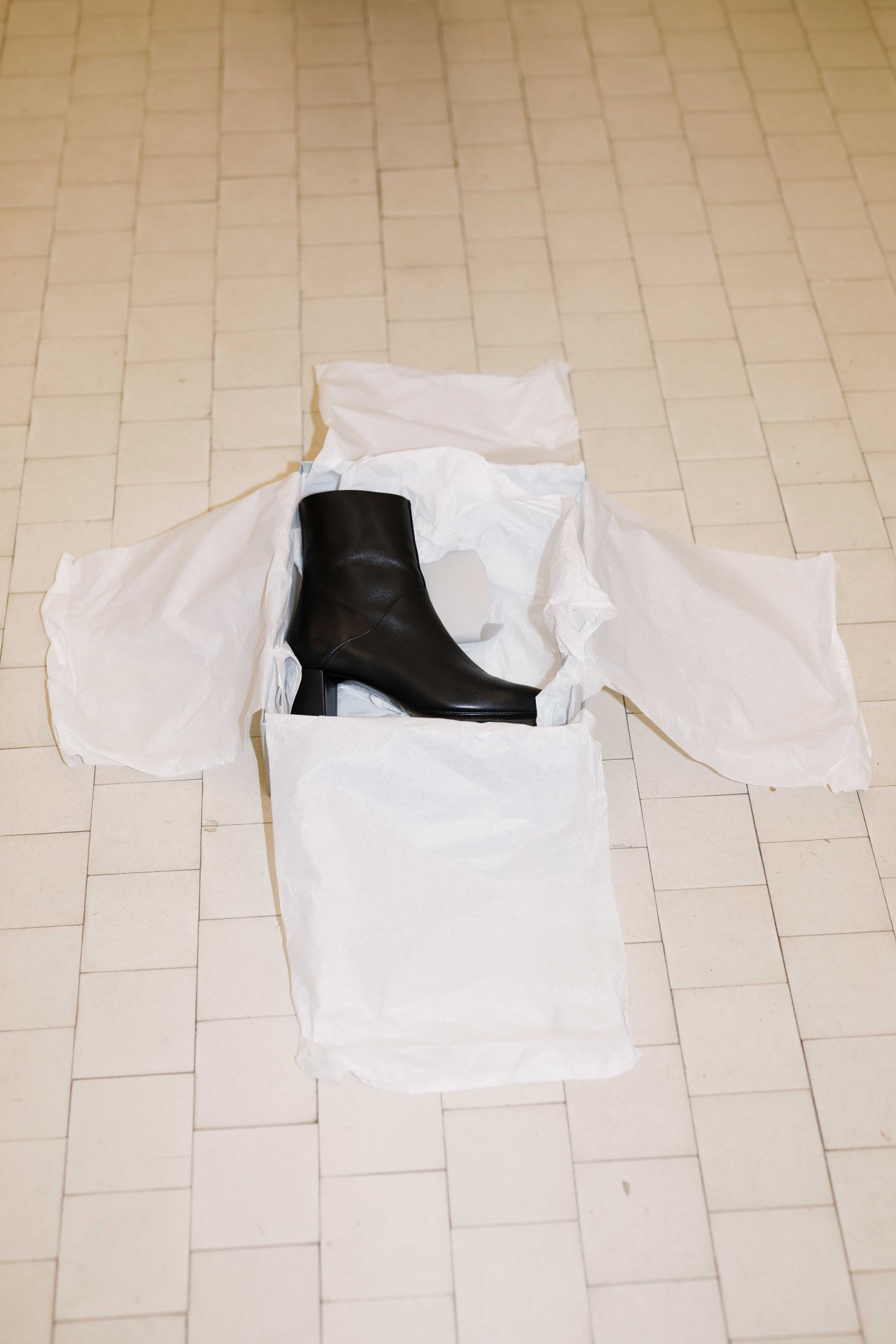 Black leather ankle boot by Anne Thomas displayed in white tissue paper inside an open white box on a tiled floor.