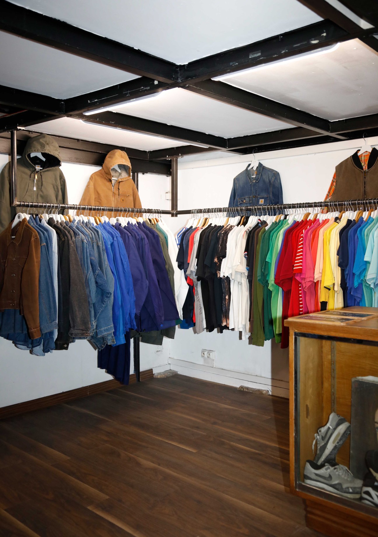 Clothing displayed on wooden racks inside a bright retail space with black ceiling beams and white panels.