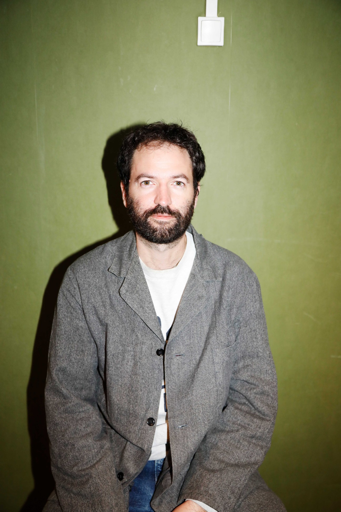 Man with dark hair and beard wearing gray blazer and white t-shirt against green wall.