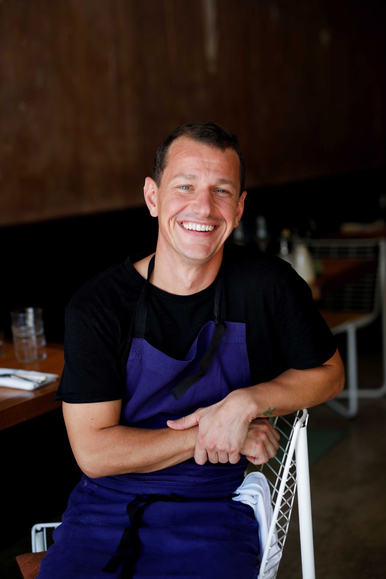 Chef Harry Cummins smiling in black shirt and blue apron in restaurant kitchen