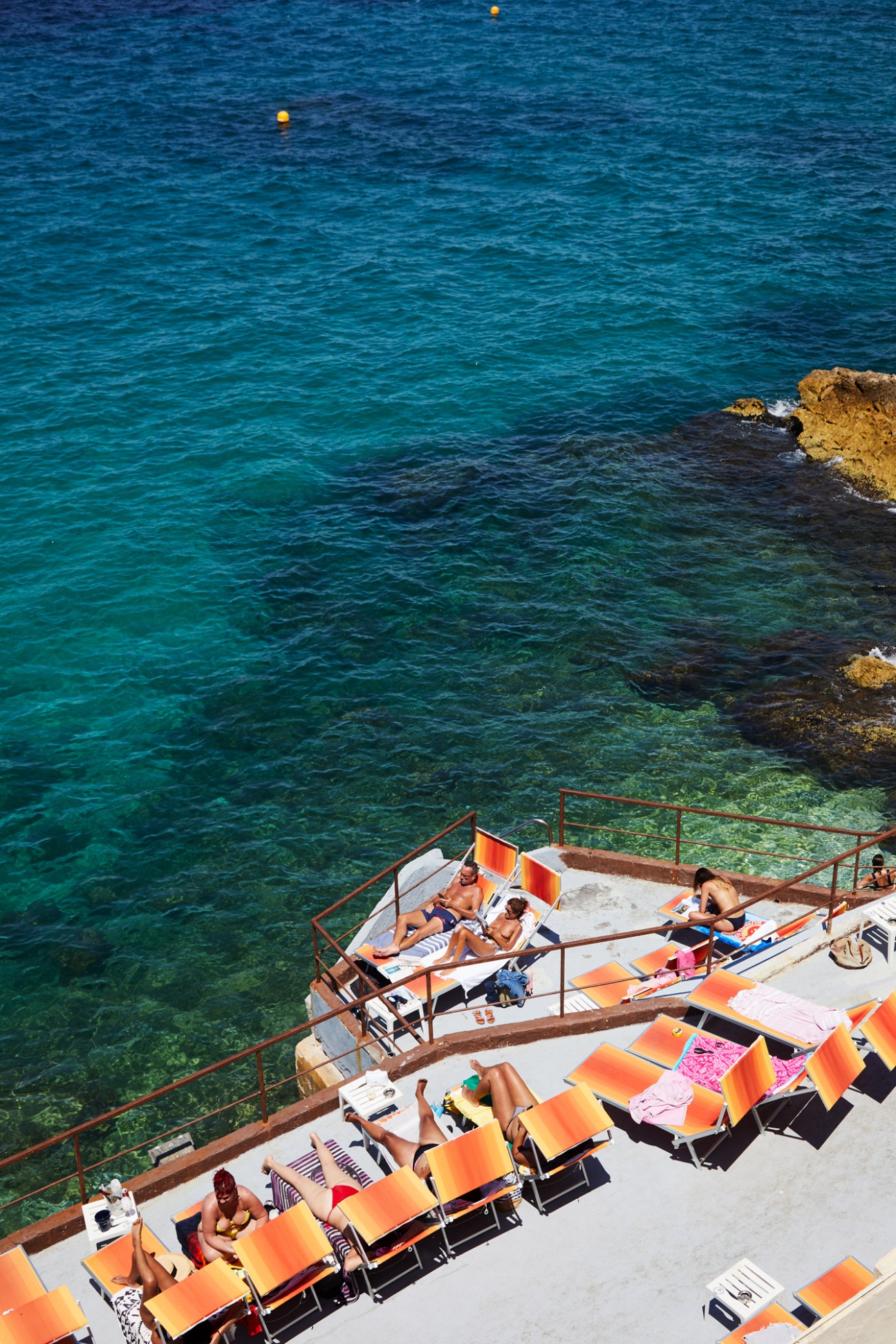 People sunbathing on orange loungers on a white concrete terrace overlooking bright blue Mediterranean water
