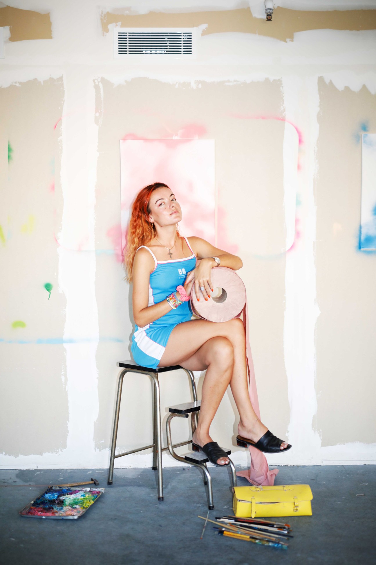 Woman in blue dress sitting on stool in art studio with paint supplies and canvases on floor