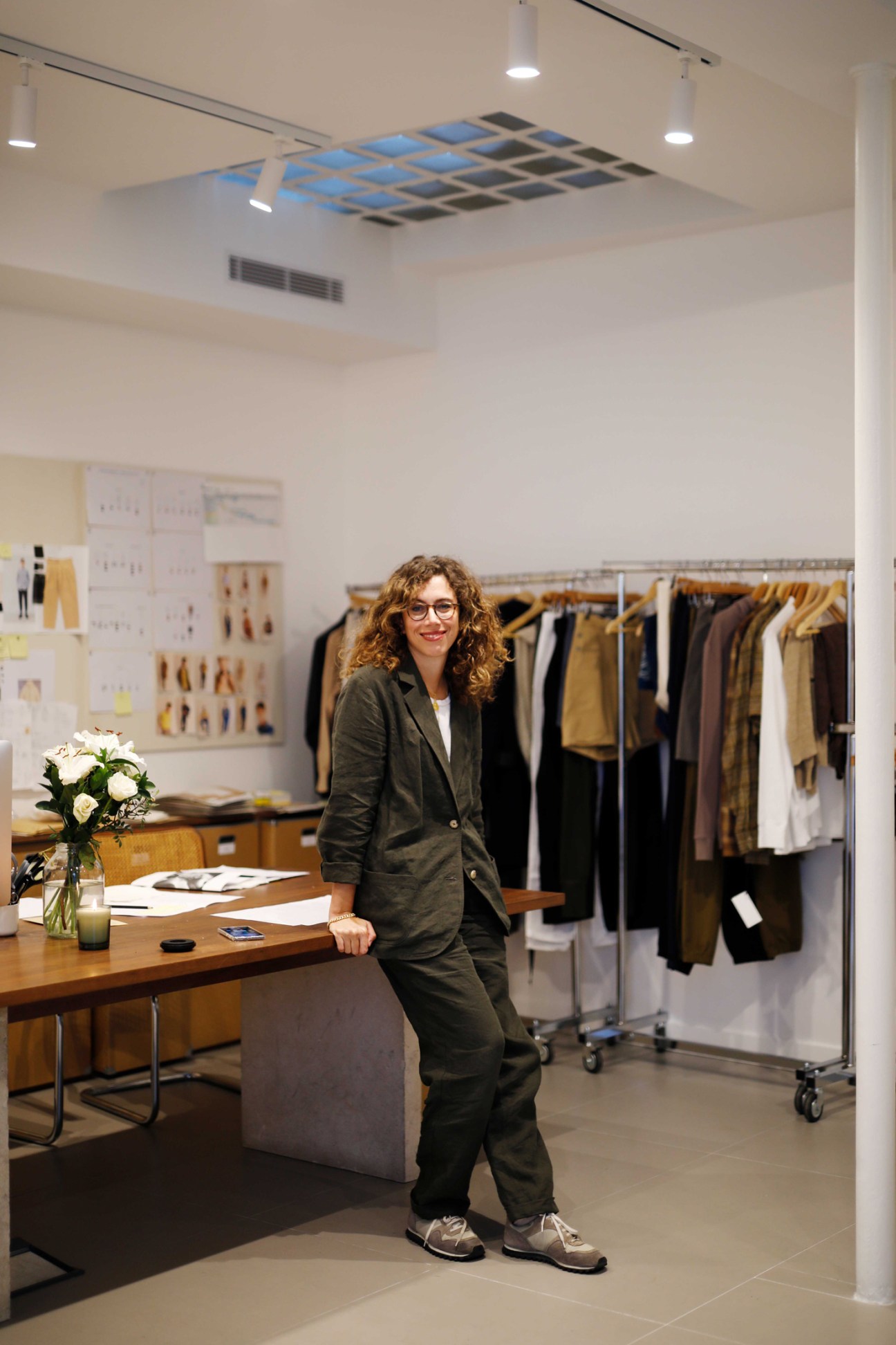 Woman in dark blazer stands in bright fashion studio with clothing racks and design samples.
