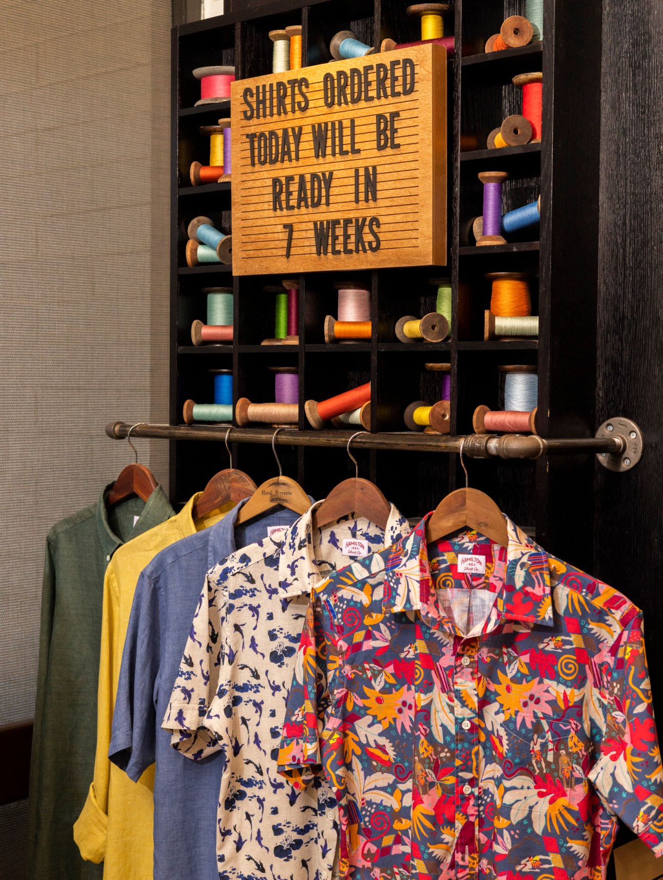 Colorful patterned shirts hanging on wooden hangers with spools of thread displayed in compartments above