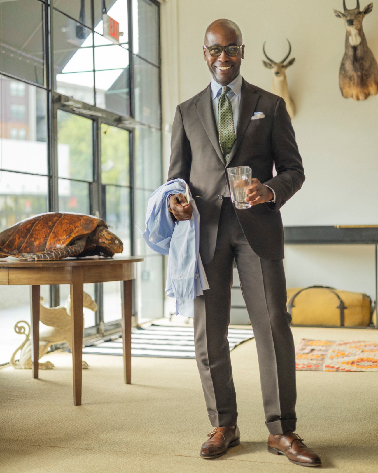 Well-dressed Black man in dark suit with green tie and glasses holding a drink in an upscale interior