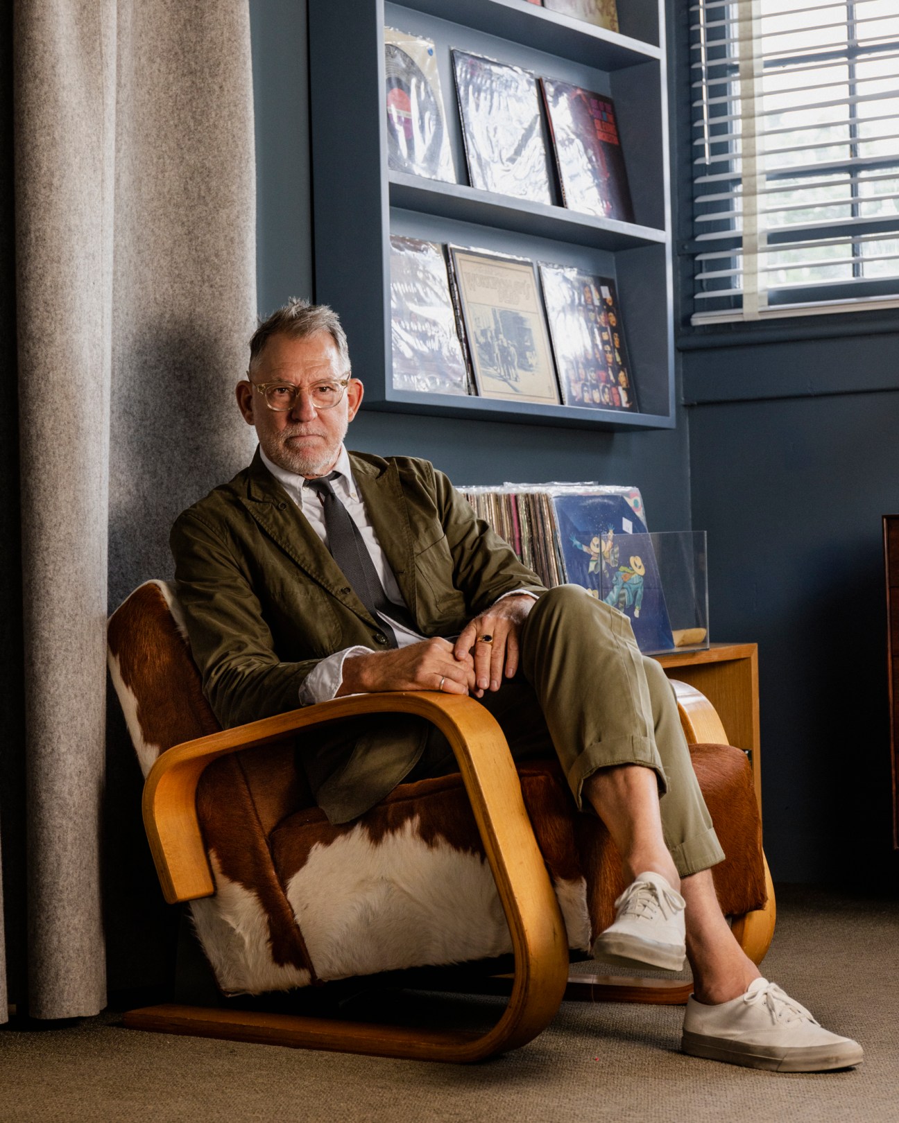 Scott Newkirk sits in a modern chair wearing an olive green suit next to shelves of vinyl records
