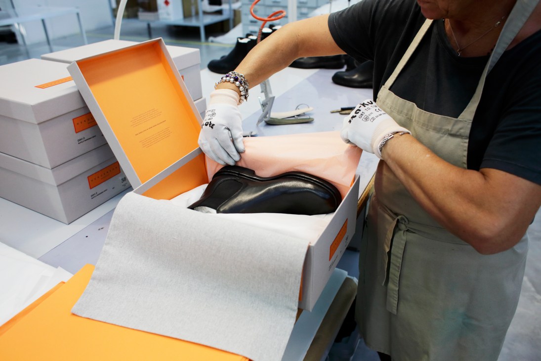 Worker in white gloves carefully placing black leather shoe into orange-lined white box at Italian footwear factory