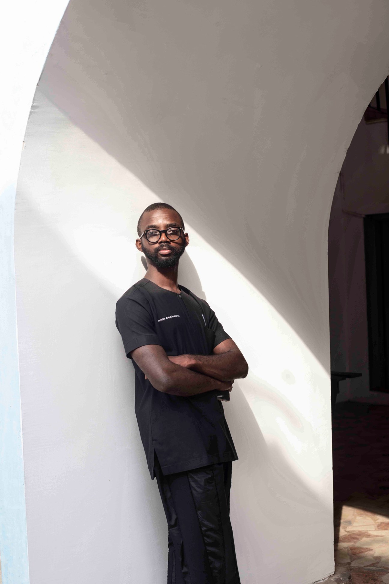 Man in black t-shirt and glasses standing against curved white wall with arms crossed.