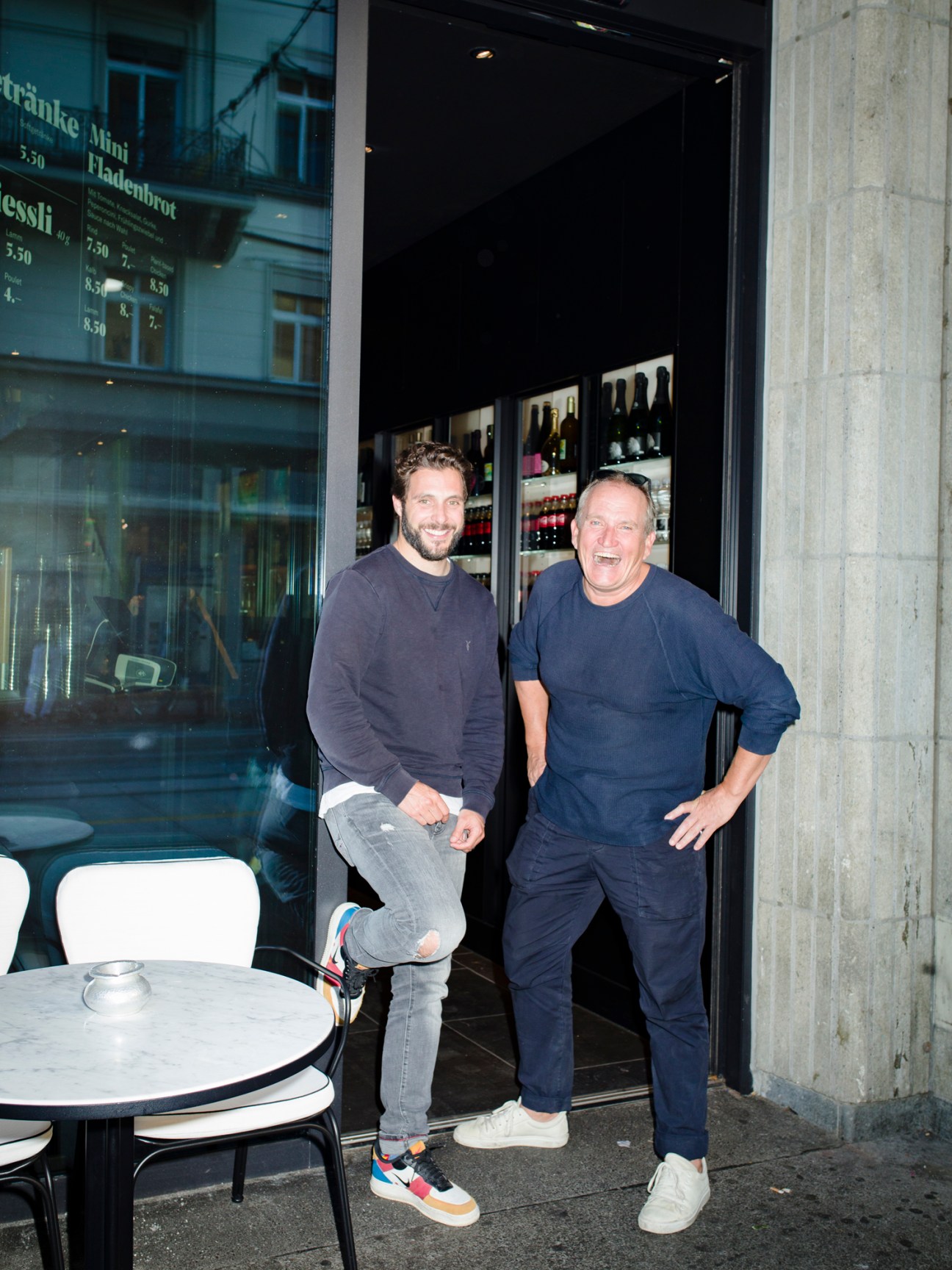 Two men standing outside nzz am Bellevue restaurant entrance, smiling at camera