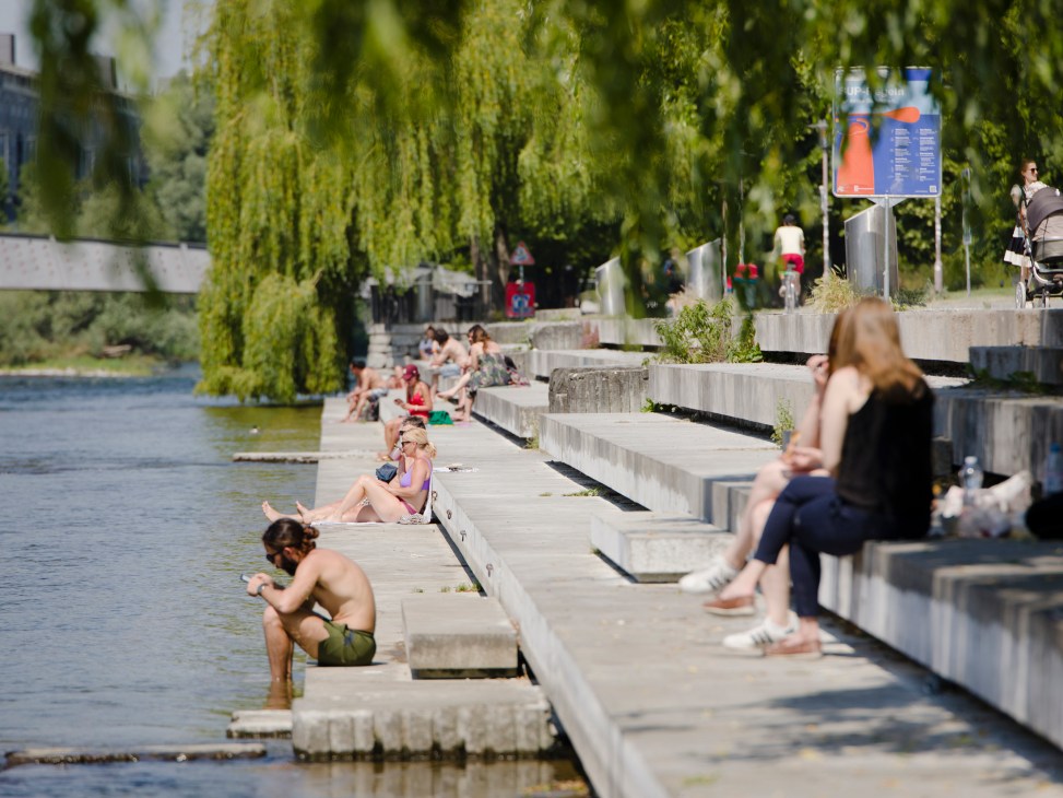 People relaxing by a waterside promenade with weeping willows and concrete steps along a river in an urban setting