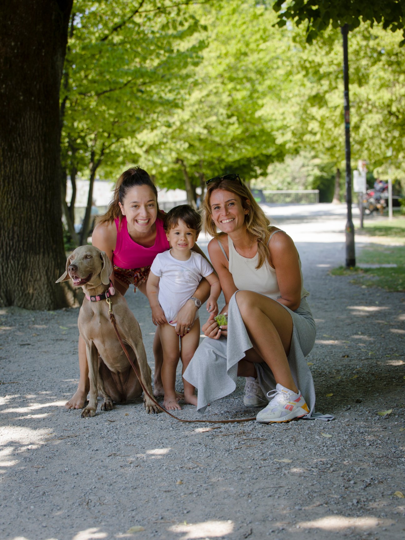Two women and a young child with a dog posing together on a tree-lined path