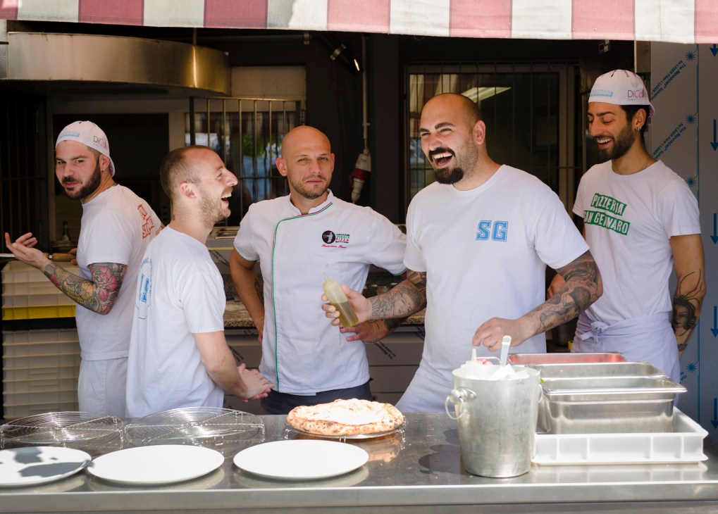 Five chefs in white uniforms and caps laughing together in a restaurant kitchen with pizza and cooking equipment