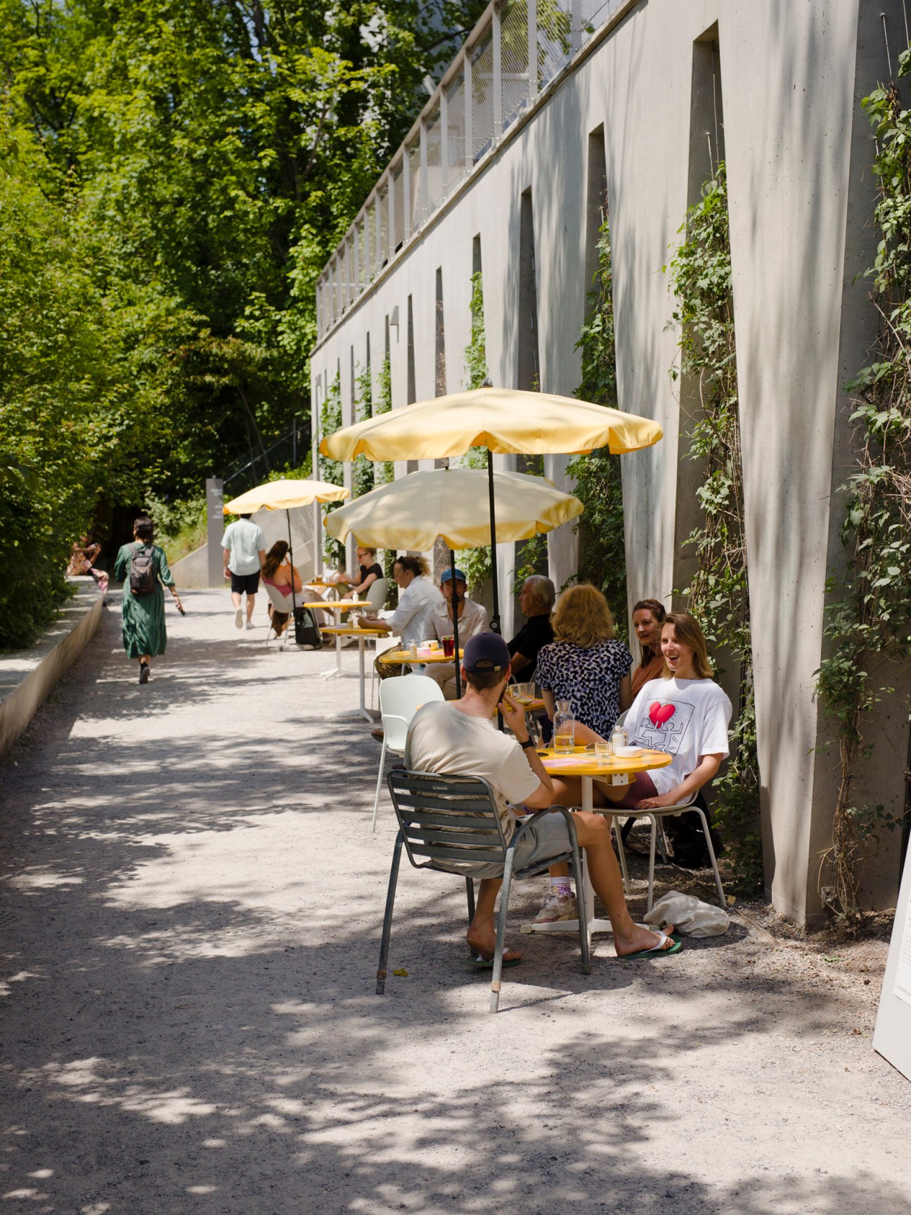 People dining at outdoor cafe tables with yellow umbrellas along a tree-lined walkway
