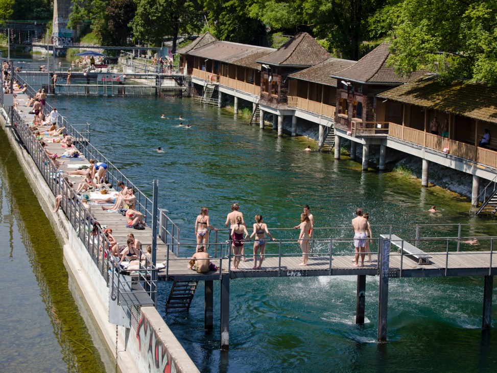 People swimming and sunbathing at a riverside lido with wooden boathouses and tree-lined banks