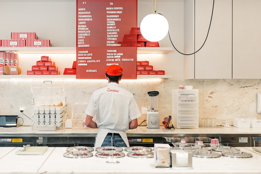 Santini gelato shop worker in red cap and white shirt standing behind counter with flavor menu board.